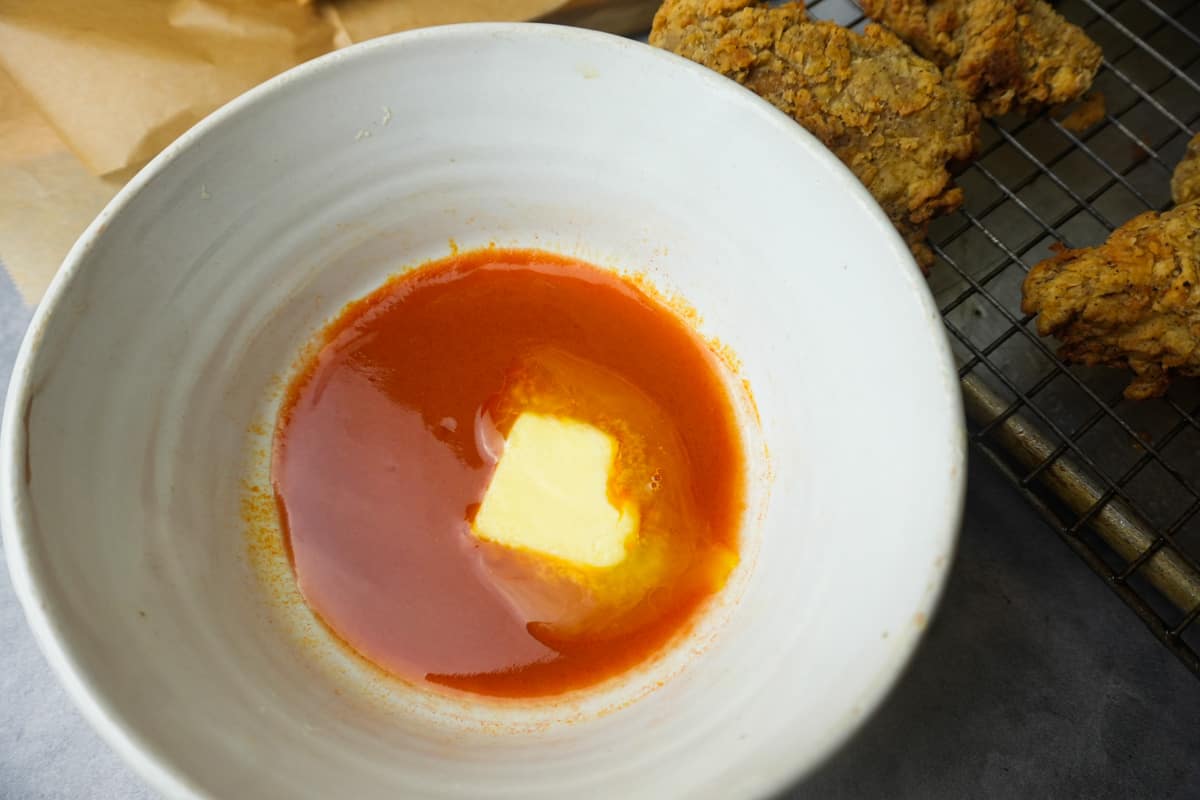a white bowl with melted vegan butter and hot sauce. Vegan fried chicken on a wire rack in the background awaiting dipping.