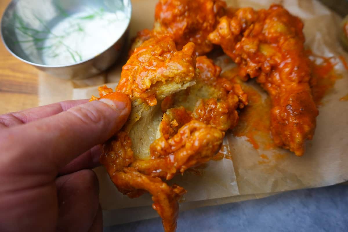 a close-up of a handheld piece of ripped-into buffalo seitan. Ranch dip and extra pieces on brown paper in the background.