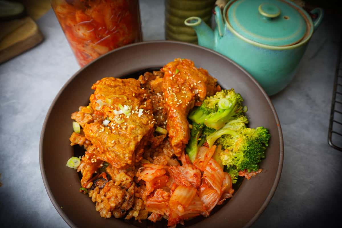 Vegan Korean Fried Chicken served with kimchi fried rice, kimchi and steamed broccoli in a brown bowl. Tea cup and pitcher in the background
