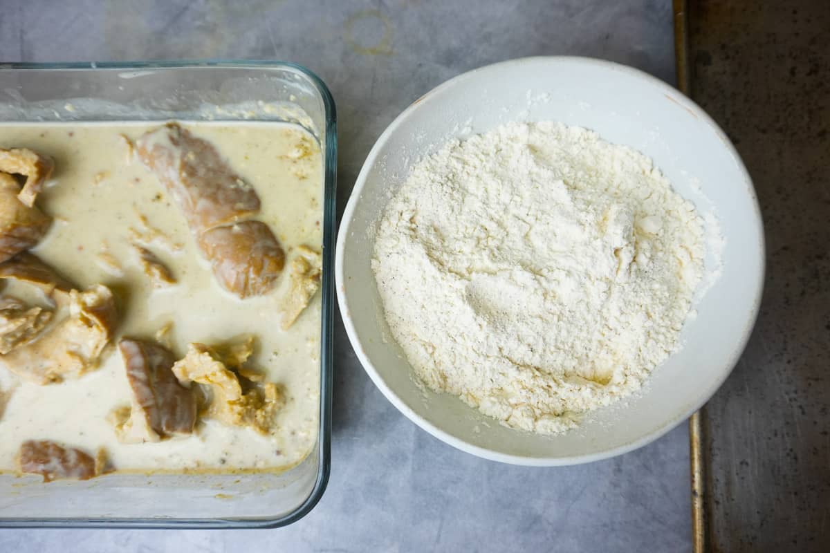 Seasoned flour mixture in a bowl with the marinated seitan in a container next to it on a concrete counter.