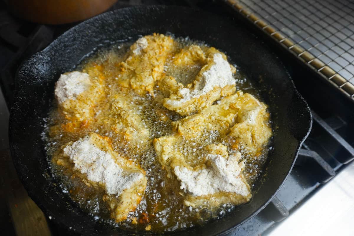 Seitan being fried in a cast iron skillet. You can see the oil bubbling and the edges of the seitan browning lightly.