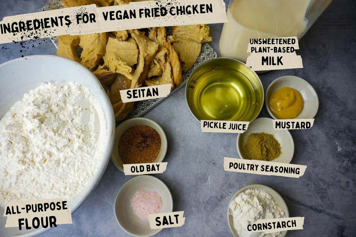All of the ingredients for vegan fried chicken measured out in bowls on a concrete counter. the seitan pieces are on a plate and the plant-based milk is in a milk bottle.