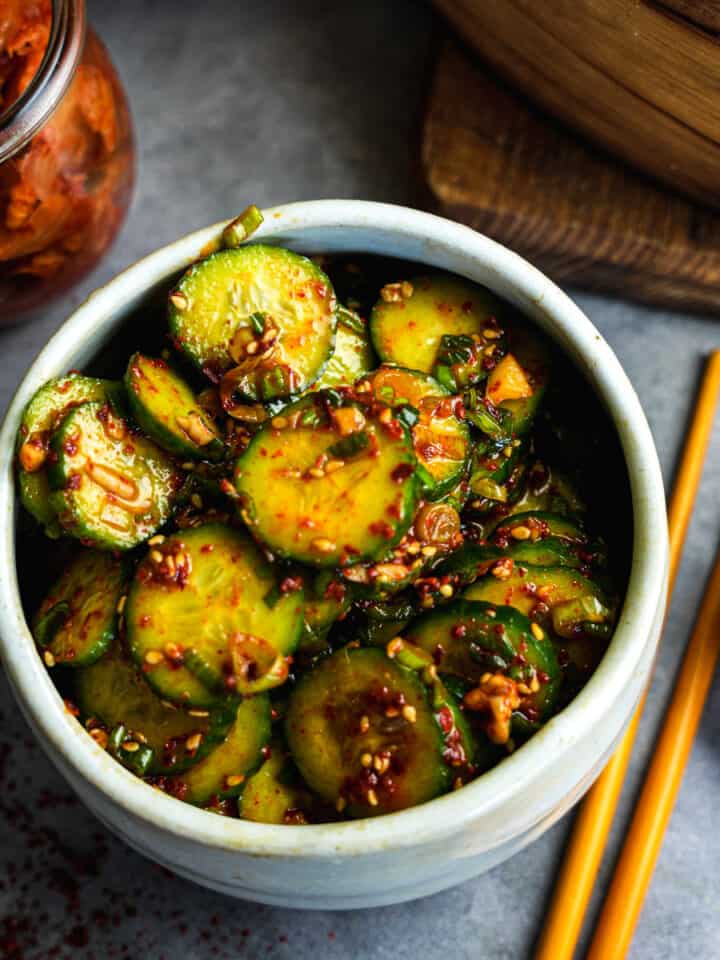 Korean cucumber salad in a white bowl with chopsticks and kimchi on the side.