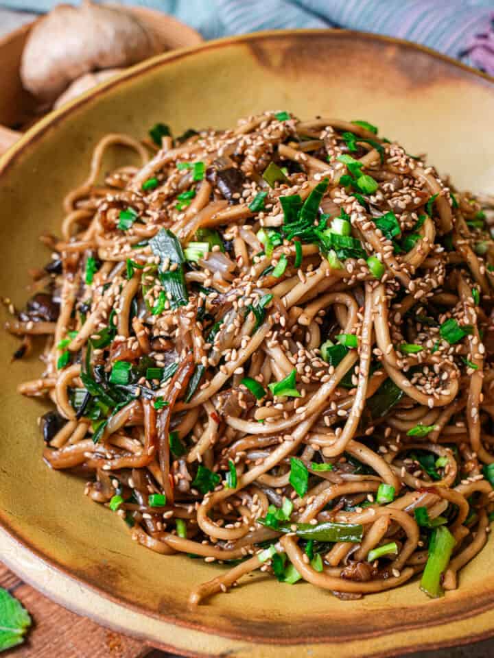 A brown handmade wide bowl with Mi Xao in it, garnished with sesame seeds and fresh chives.