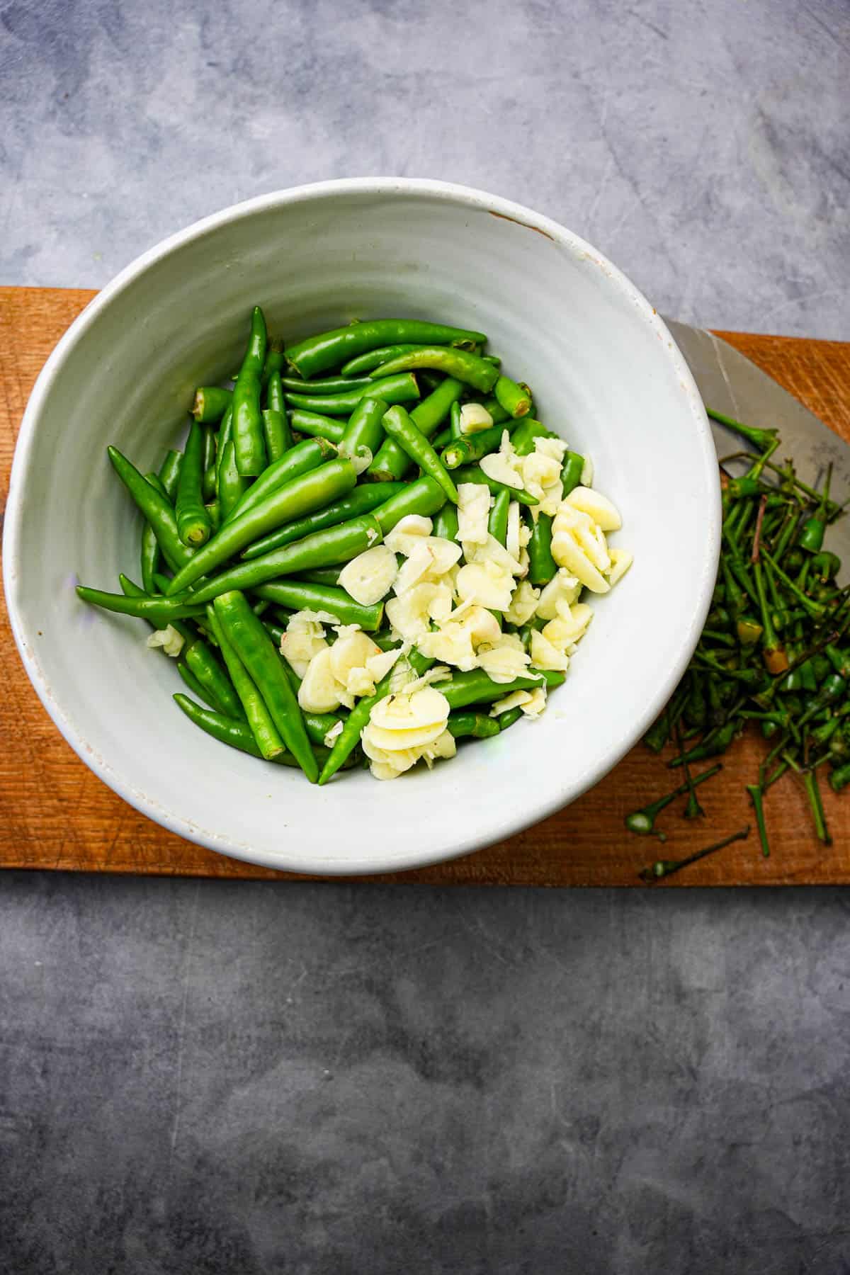 green chilies and garlic in a white bowl on a cutting board. Chili stems are on the side.