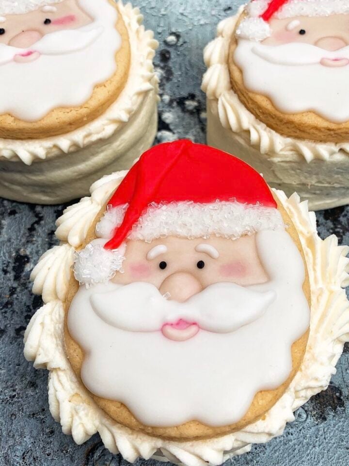 Three vegan Christmas cookies decorated with Santa Claus faces featuring white icing for the beard and red icing for the hat, placed on a rustic gray surface.