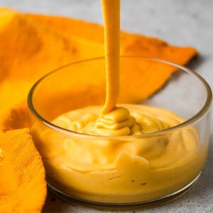 A thick, creamy yellow plant-based cheese sauce being poured into a transparent glass bowl, with an orange cloth partially visible beside it.