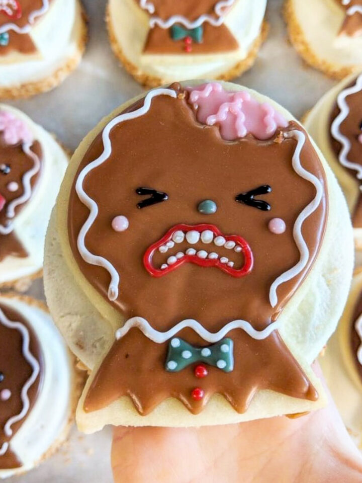 A hand holds a decorated, vegan cookie featuring a surprised or distressed gingerbread person with icing details. More similarly decorated vegan cookies are in the background.