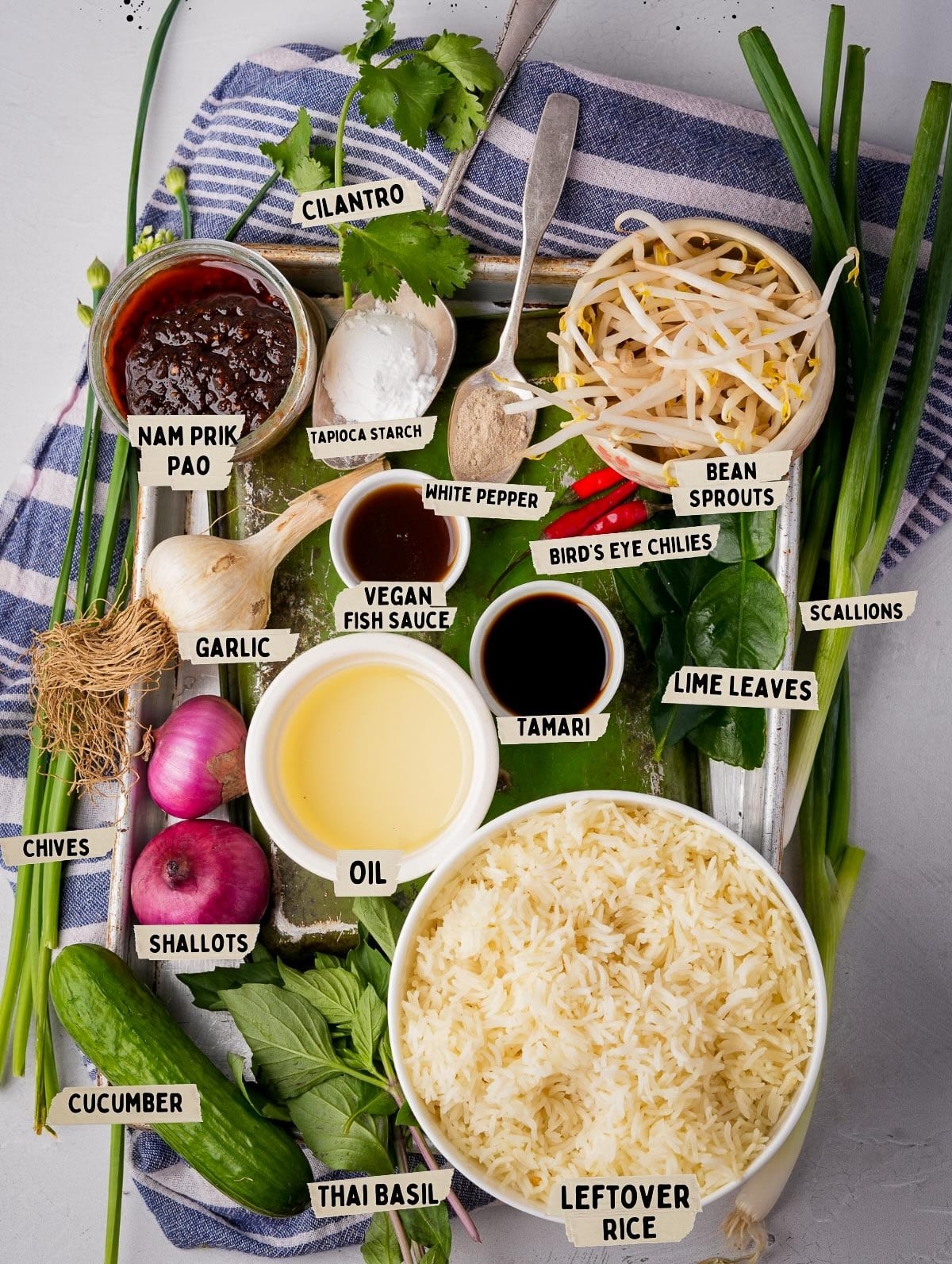 A variety of ingredients for Khao Pad on a table, including cilantro, bean sprouts, leftover rice, garlic, shallots, and seasonings like nam prik pao, vegan fish sauce, and tamari.