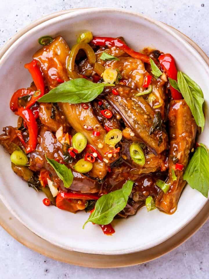 A bowl of stir fried Thai basil eggplant garnished with chili pepper slices, thai basil leaves and scallions in a bowl with a fork.