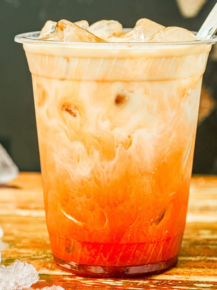 A plastic cup filled with iced Thai tea, showing a gradient of red tea and milk, sits on a wooden surface with ice cubes scattered around-a delightful snapshot of the perfect Thai tea recipe.