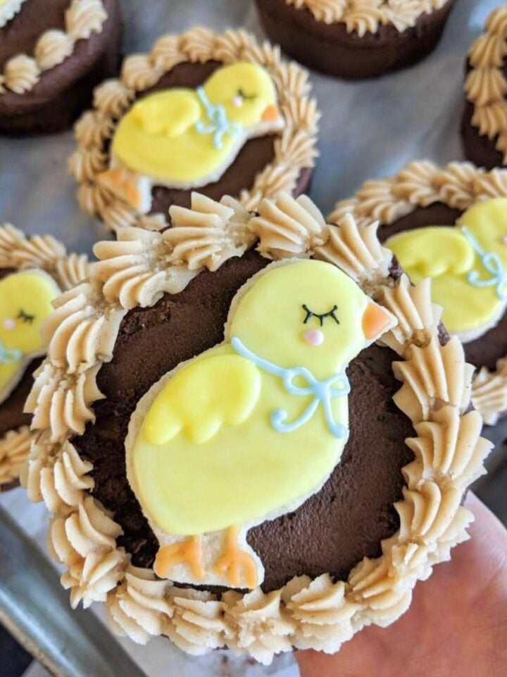 A hand holds a round vegan chocolate cupcake topped with a frosting design of a yellow chick wearing a blue ribbon. More cupcakes with similar Easter designs are visible in the background.