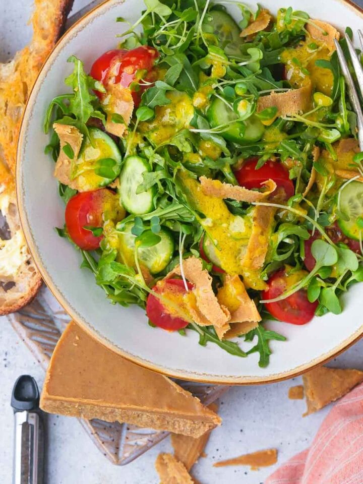 A healthy salad with arugula, tomatoes, cucumbers, Vegan Parmesan Cheese, and a creamy dressing, served with a side of toasted bread.