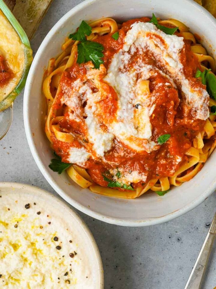 A bowl of fettuccine pasta topped with tomato sauce and a dollop of vegan ricotta cheese, garnished with basil leaves.