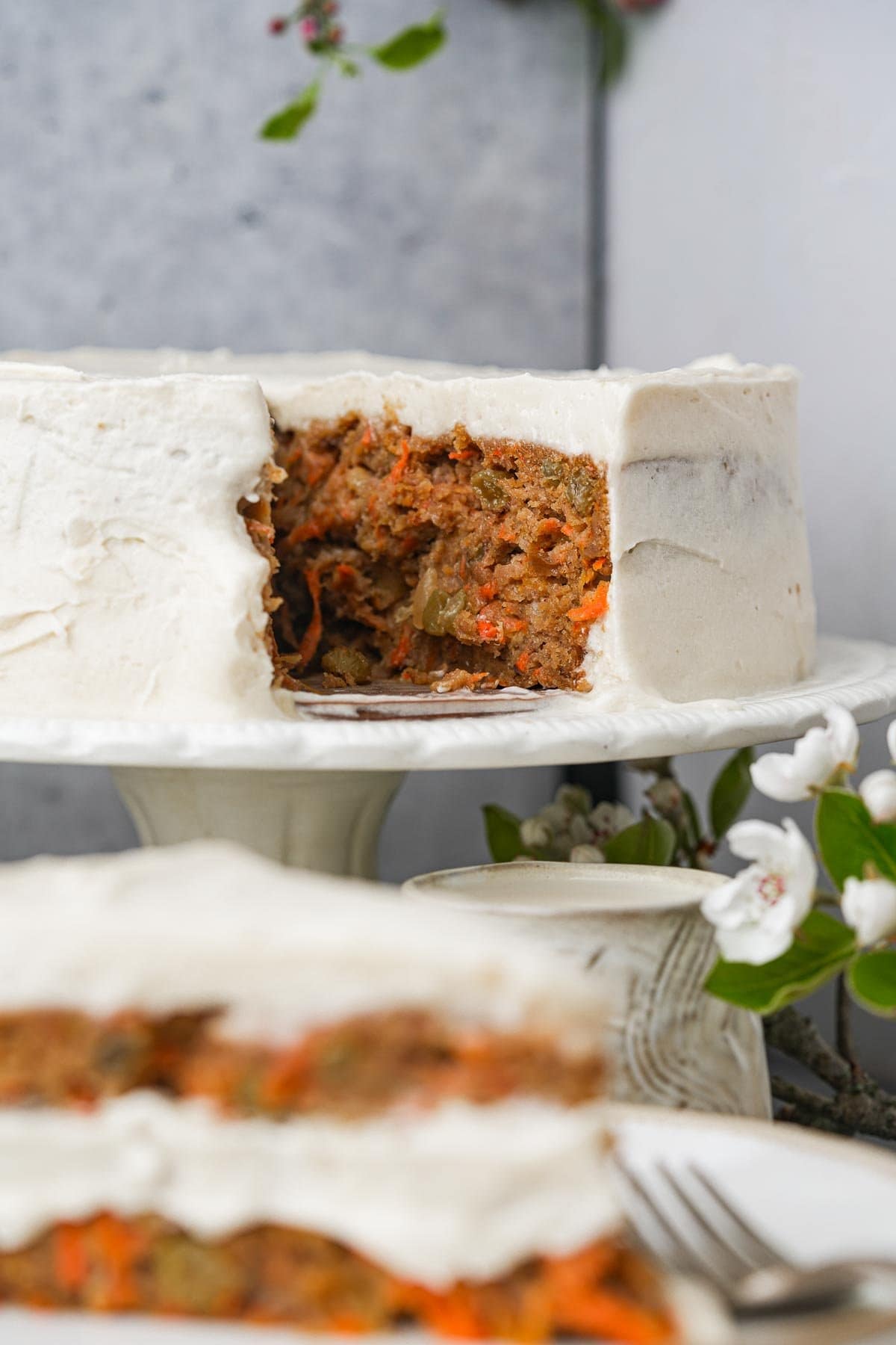 A vegan carrot cake with a slice removed, displayed on a white cake stand with cream cheese frosting, surrounded by flowers.