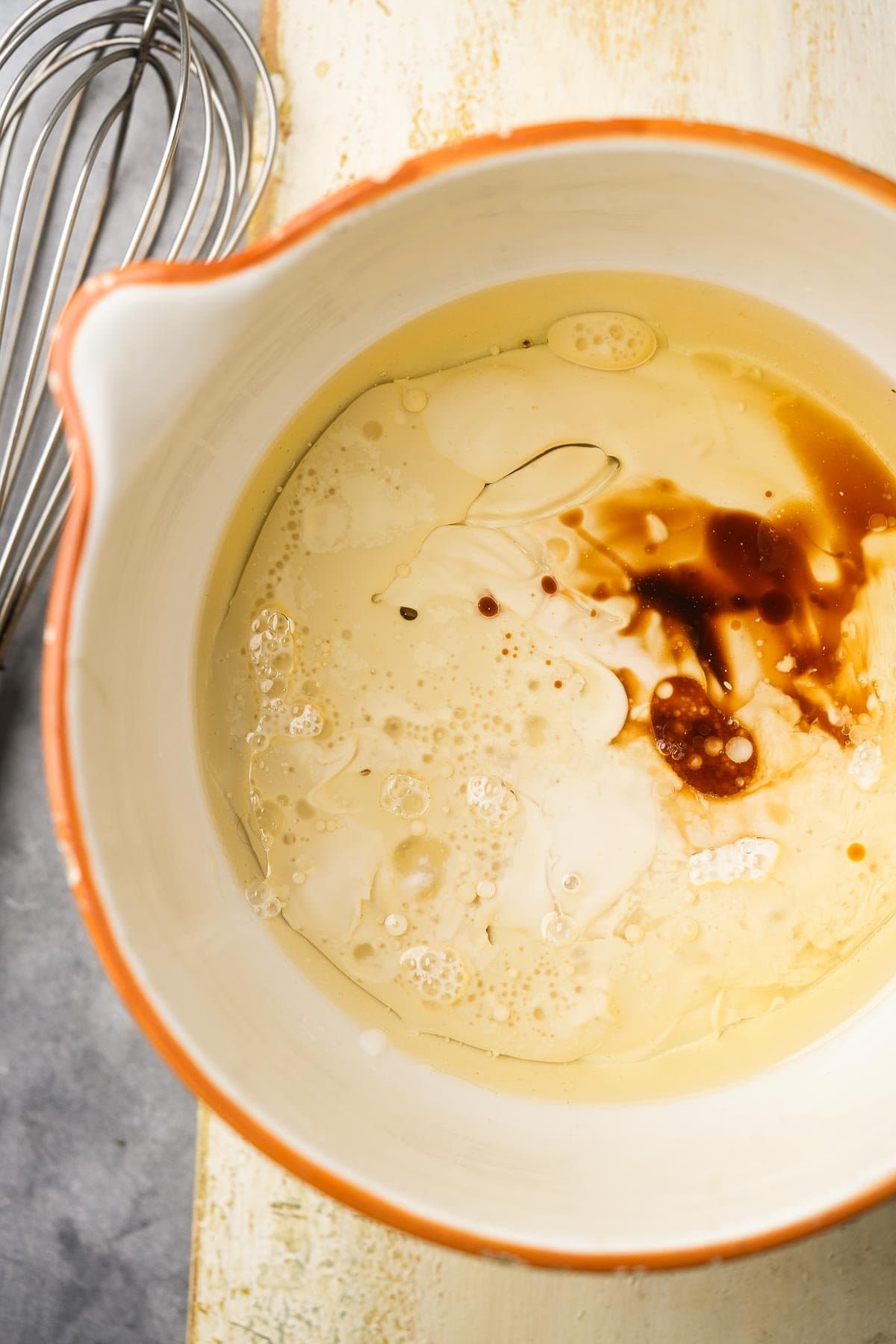 Bowl with wet ingredients for vegan carrot cake, next to a metal whisk on a light surface.