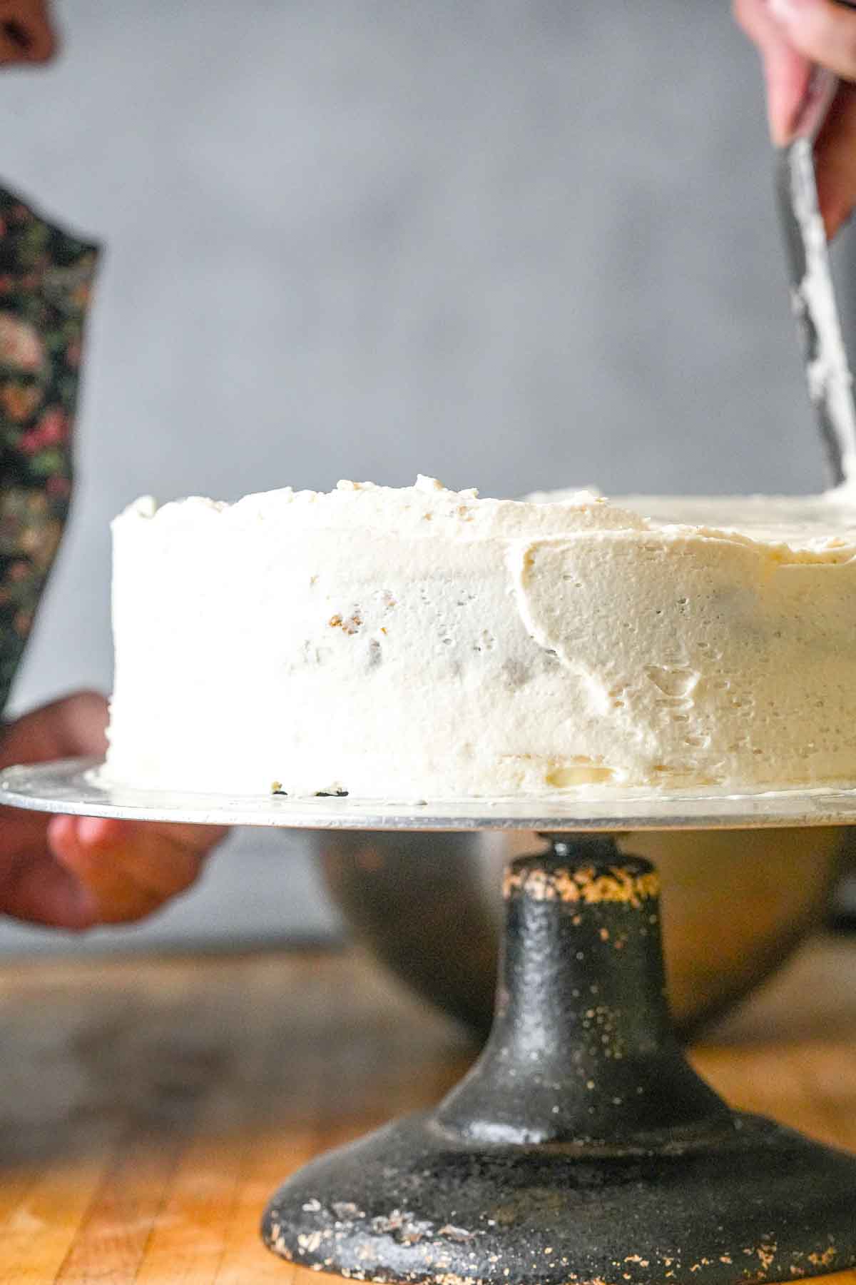 A person icing a white cake on a black cake stand, set on a wooden counter.