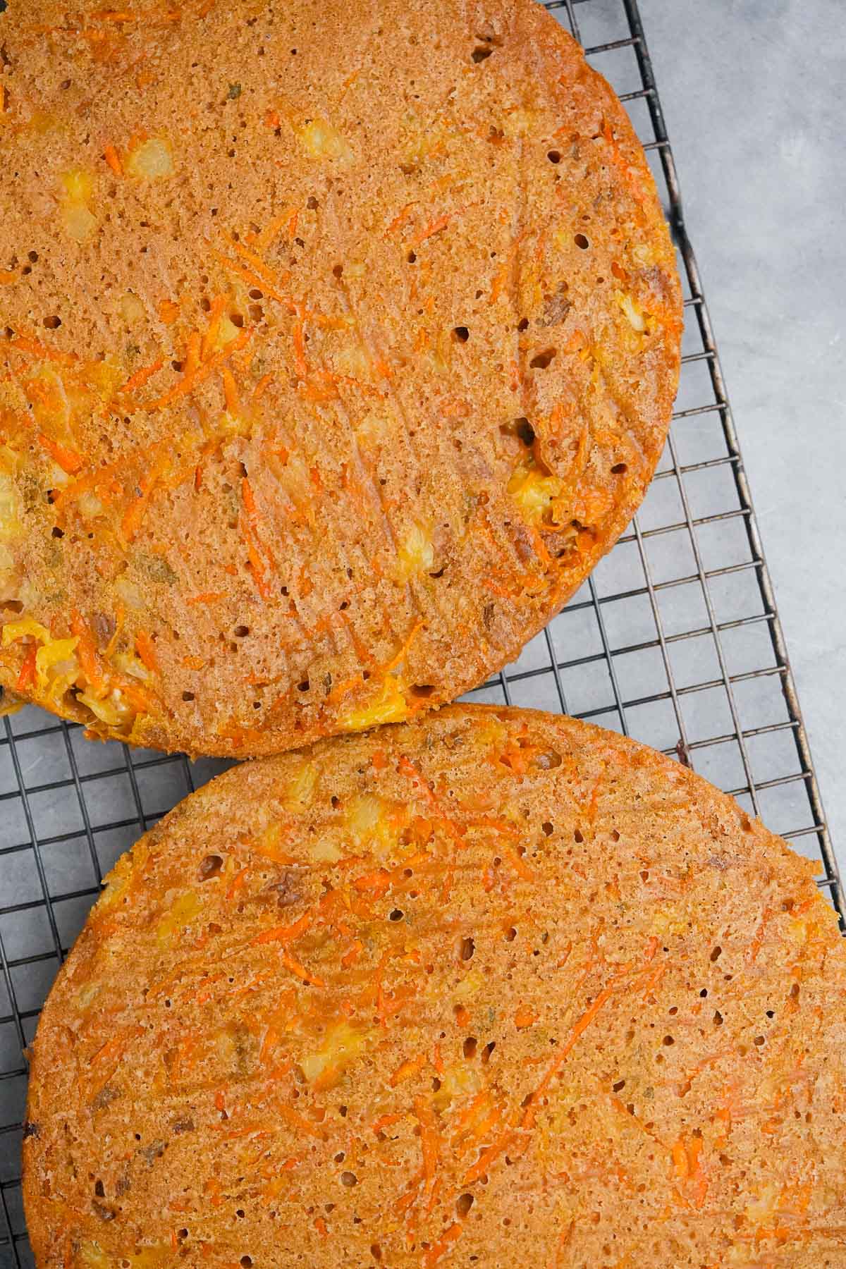 Two carrot cakes cooling on a wire rack, one partially visible, with visible carrot shreds and a golden-brown crust.