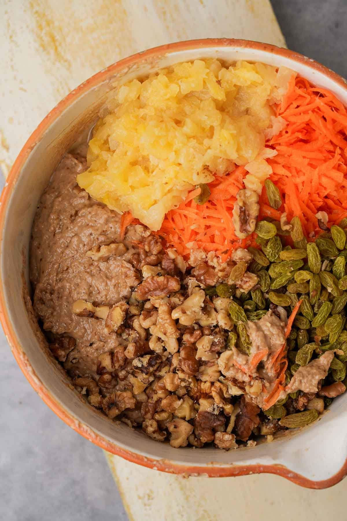 A bowl of food with sections of mixed vegan carrot cake batter, shredded carrots, chopped nuts, pineapple, and green raisins.
