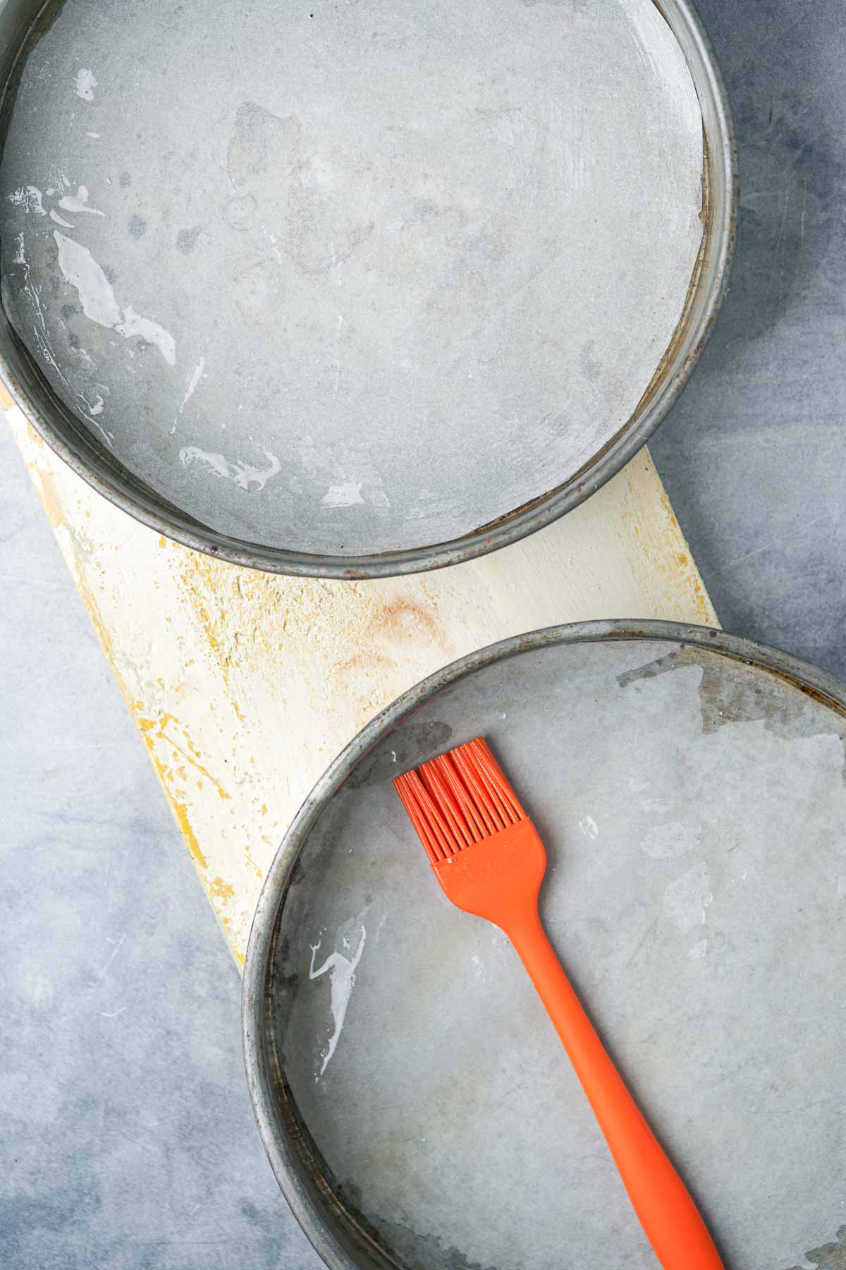 Two lightly oiled, parchment paper lined cake pans with an orange pastry brush on a kitchen counter.