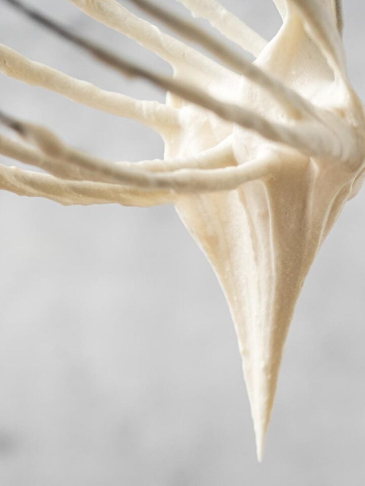 Whisk with Vegan cream cheese frosting dripping off, against a blurred light background.