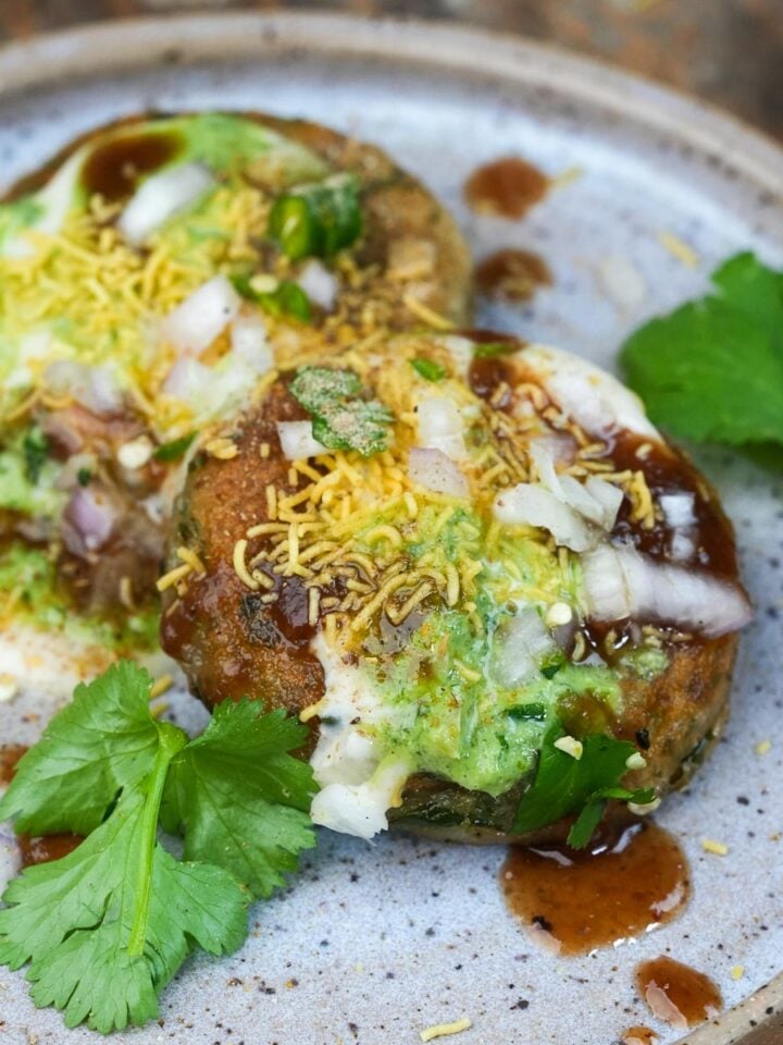 A plate of two aloo tikki chaat pieces topped with yogurt, tamarind chutney, green chutney, sev, chopped onions, and garnished with fresh cilantro leaves.