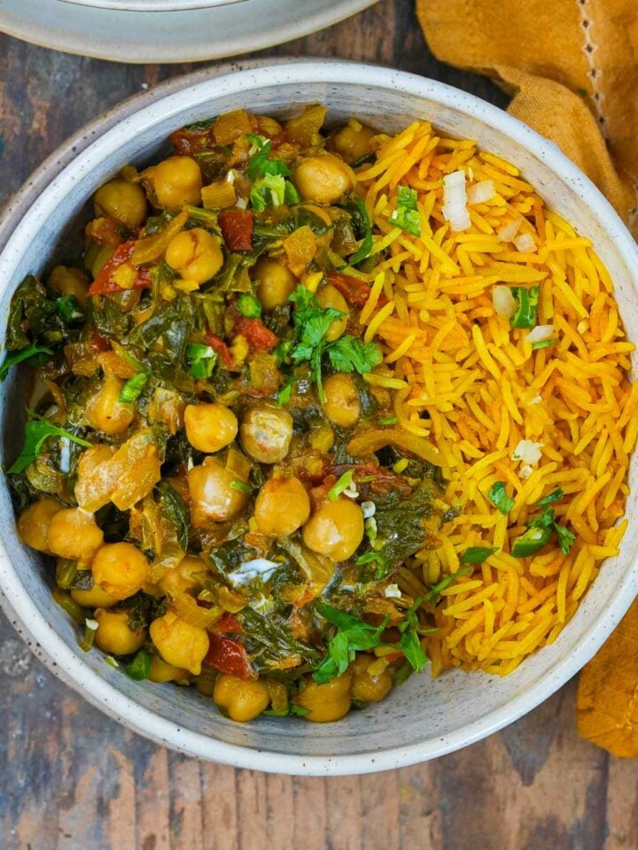 A bowl containing yellow rice and chickpea spinach curry, placed on a wooden surface beside a yellow napkin and a fork.