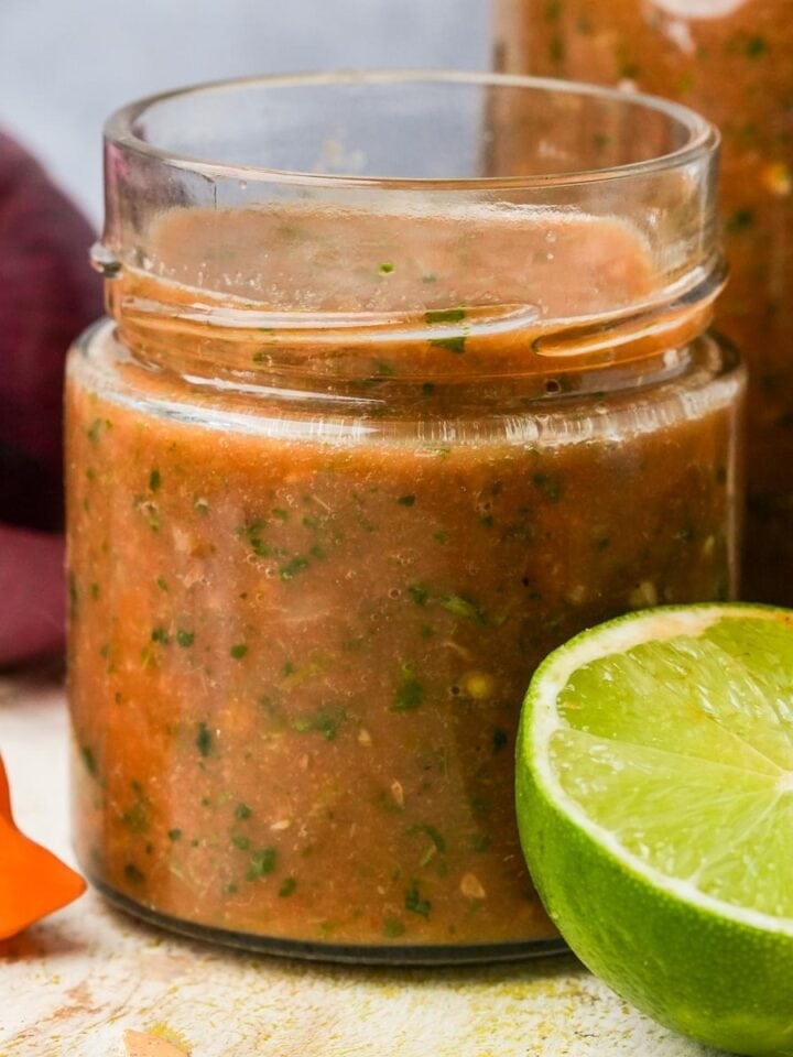 A glass jar filled with homemade habanero salsa, accompanied by a lime slice and a whole tomato, set on a rustic kitchen countertop.