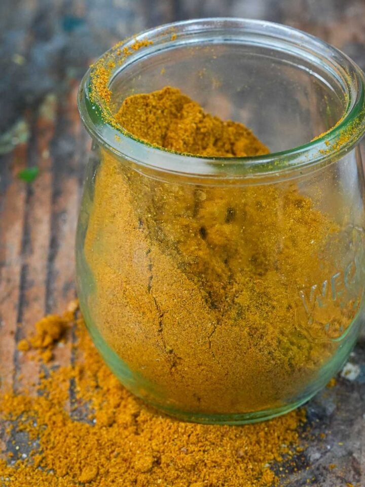 A glass jar filled with yellow Madras Curry Powder, some of which is spilled on the wooden surface.