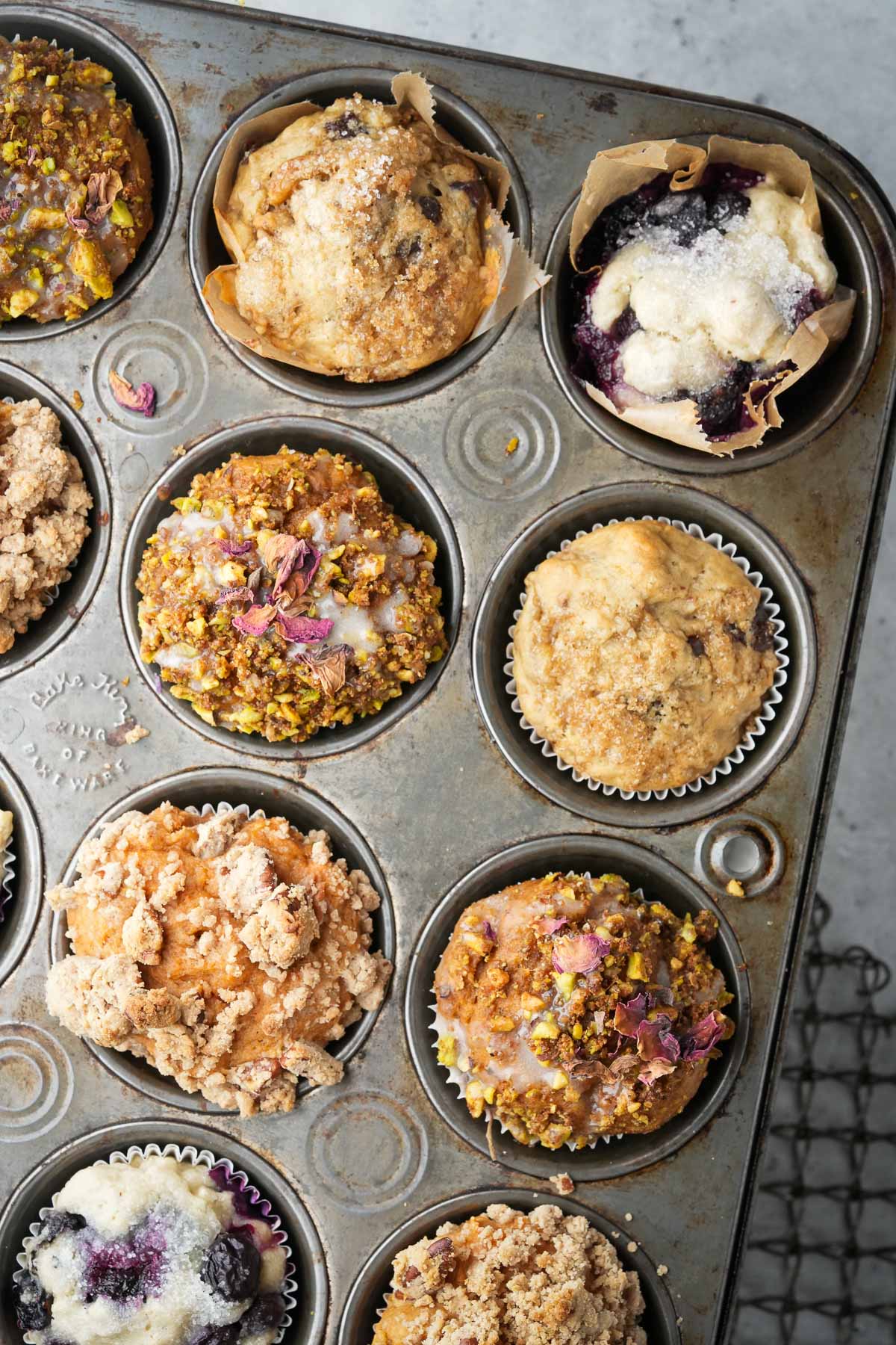 A top-view image of a muffin tin containing nine assorted muffins with various toppings, including streusel, nuts, and icing. The muffins appear freshly baked and are placed on a cooling rack.