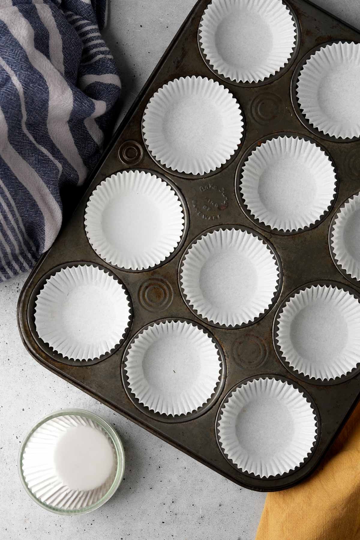 A muffin tin lined with white paper baking cups, placed on a gray surface. A folded blue and white cloth is on the left side, and a yellow cloth is on the bottom right next to a small bowl with white liquid.