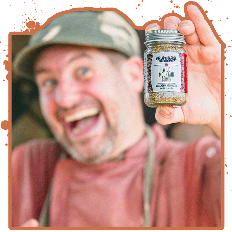 A person in a cap and apron holds up a jar labeled "Wild Mountain Cumin, Burlap & Barrel." The person is smiling and appears excited.
