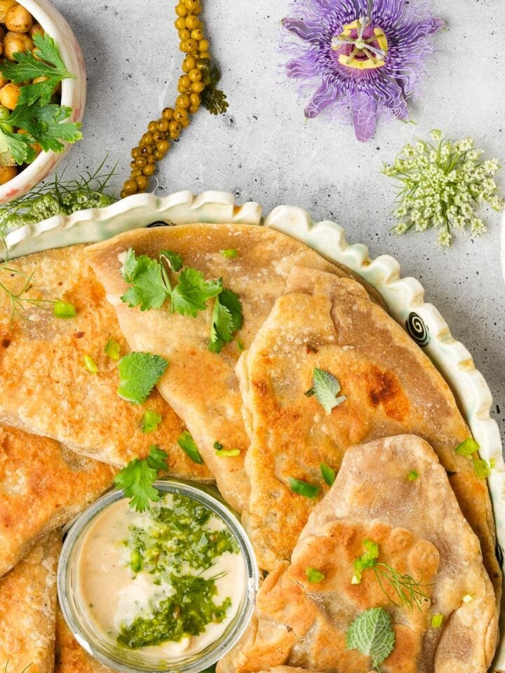 A plate of naan bread with a small dish of creamy dip, surrounded by a bowl of seasoned chickpeas, a flower, and leafy herbs on a light-colored surface.