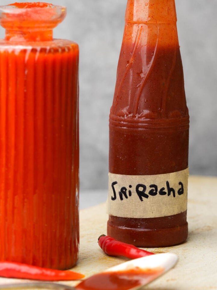 Two bottles of red hot sauce sit on a surface. One bottle is labelled "Sriracha," and a spoon with sauce and a small red chili pepper are in the foreground.
