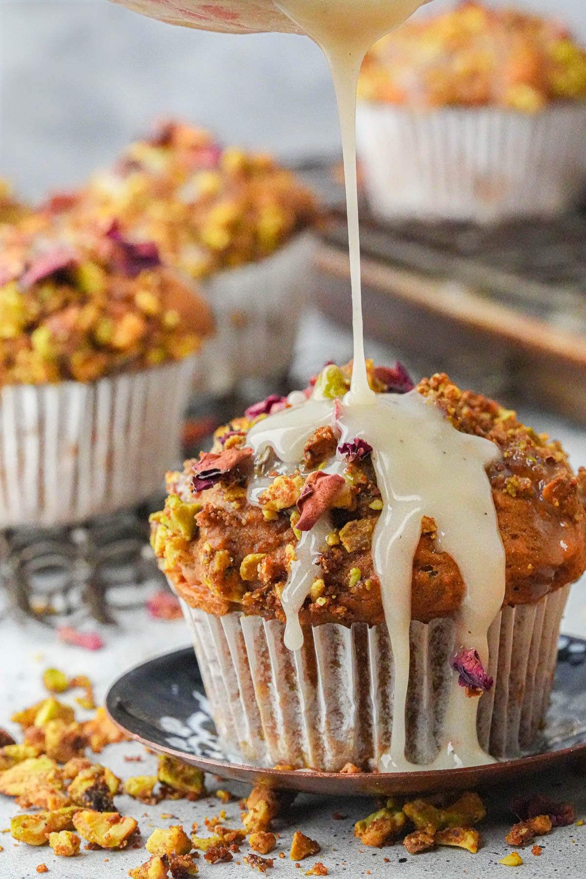 A close-up of a Vegan Pumpkin Muffin topped with crushed nuts and flower petals, with icing being drizzled over it.