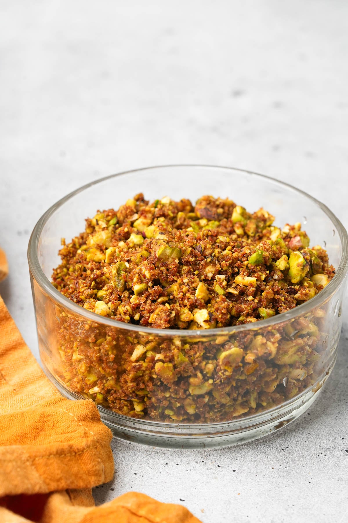 A glass bowl filled with chopped and seasoned nut mixture placed on a gray surface with an orange cloth next to it.