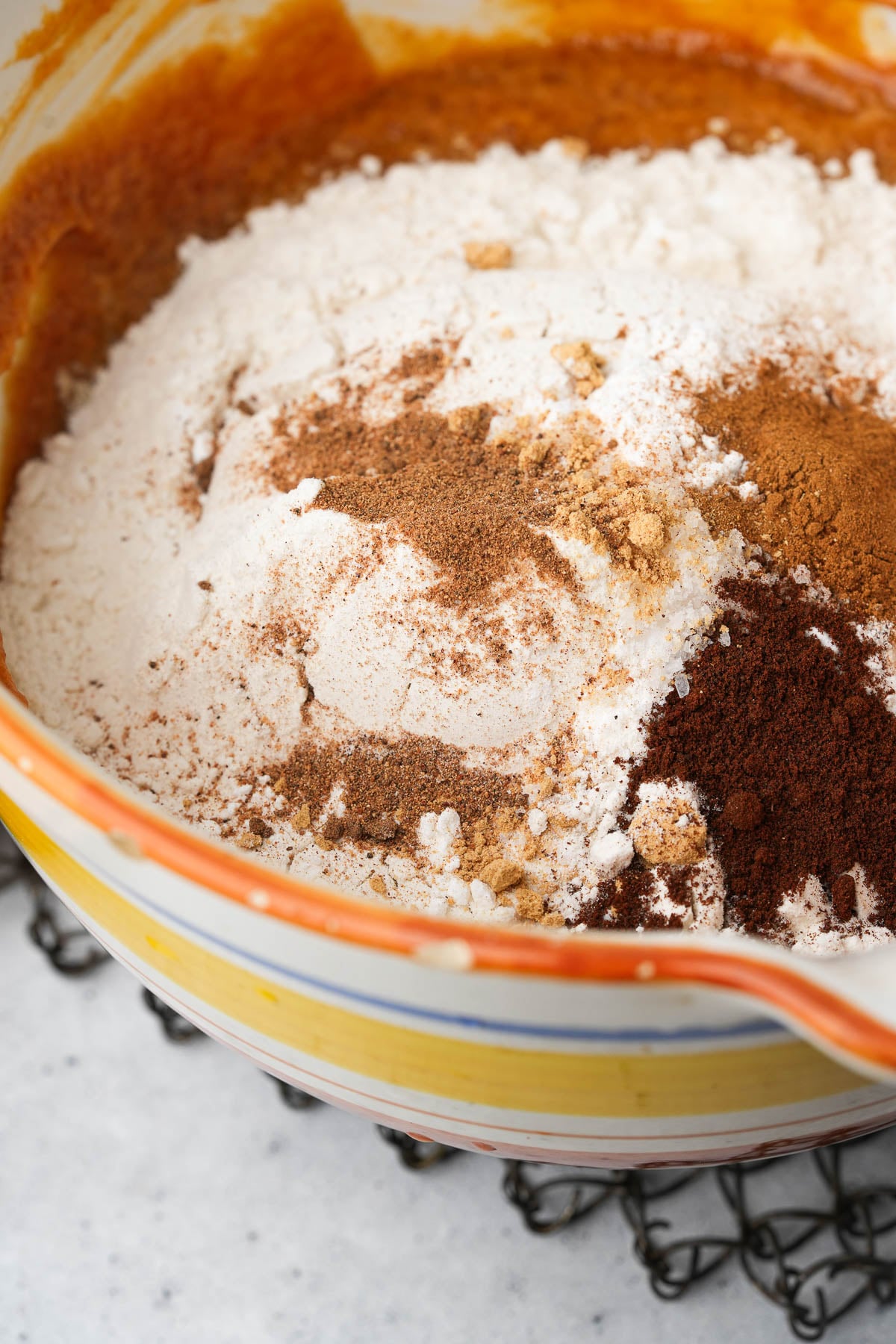 A ceramic bowl contains a mixture of dry baking ingredients, sifted on top of the wet mix to make Vegan Pumpkin Muffins.