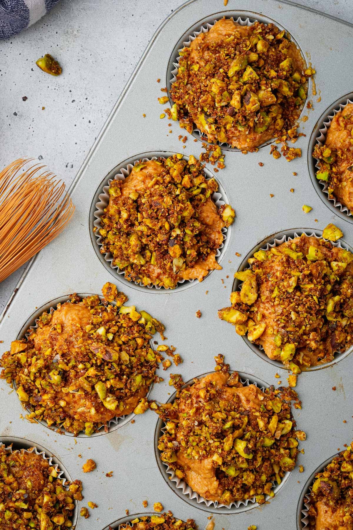 Close-up of a muffin tin with nine unbaked muffins topped with chopped nuts and streusel in paper liners on a grey surface, with a pastry brush nearby.