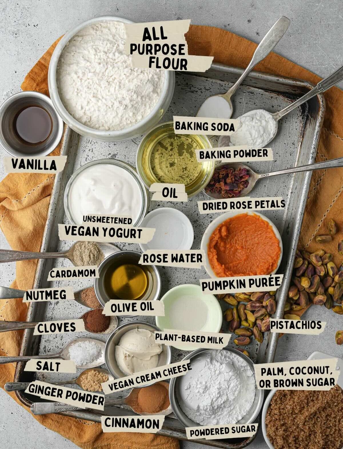 Baking ingredients arranged on a tray include all-purpose flour, vanilla, vegan yogurt, spices, oils, baking soda, baking powder, rose water, pumpkin purée for vegan pumpkin muffins, nuts, plant milk, powdered sugar, and cream cheese.