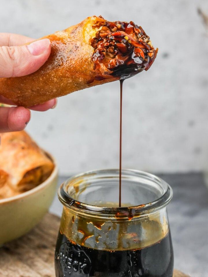 Hand dipping a spring roll into a glass jar filled with thick, dark kecap manis. A plate with more spring rolls is visible in the background.