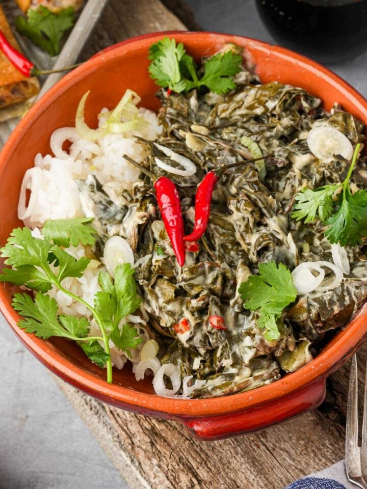 A bowl of white rice and Filipino Laing topped with red chili peppers, and cilantro, placed on a wooden surface with a fork and rolls in the background.