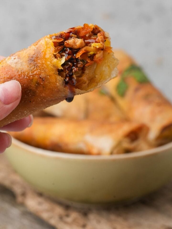 Close-up of a hand holding a Lumpiang Shanghai roll filled with vegetables and soy sauce, with more spring rolls in a bowl in the background.