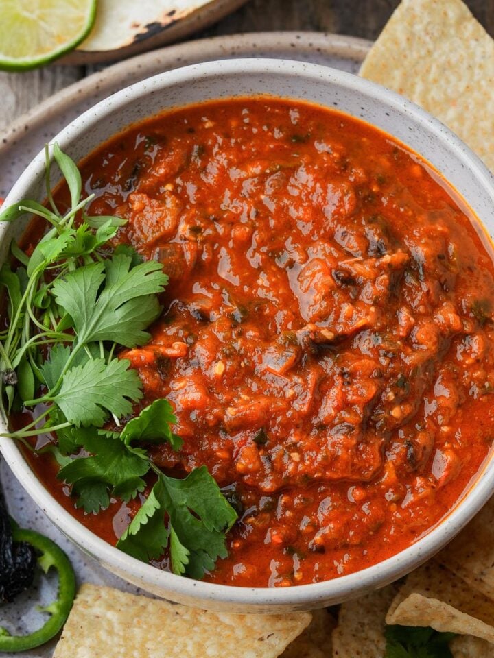 A bowl of red salsa rojo garnished with cilantro, surrounded by tortilla chips, a slice of tomato, lime wedge, and dried pepper on a wooden surface.