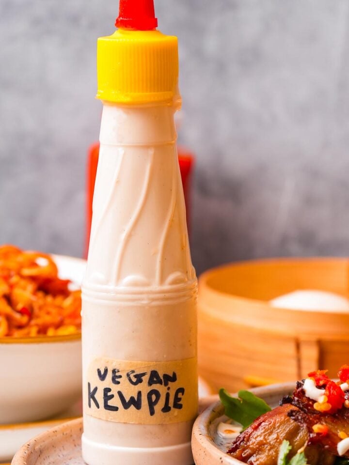 Bottle of vegan Kewpie mayonnaise with a yellow cap placed in a white bowl, surrounded by dishes of food including noodles and a serving of seitan with vegetables, set against a grey background.
