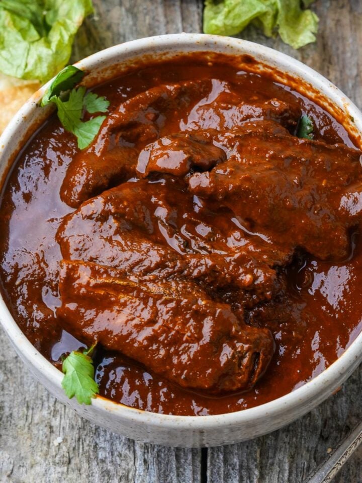 A bowl filled with red Adobo Sauce with chipotle peppers, garnished with cilantro, placed on a wooden surface near some tortilla chips and lettuce leaves.