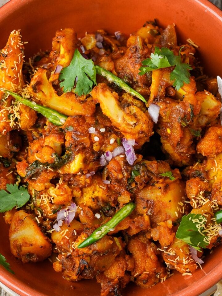 A bowl of Aloo Gobi garnished with cilantro, green chilies, chopped onions, and sev, with a fork on the side.