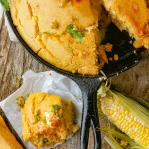 A skillet with vegan cornbread, a slice served on parchment paper next to it, and a corn cob placed on a wooden surface. The cornbread is topped with herbs and a pat of butter.