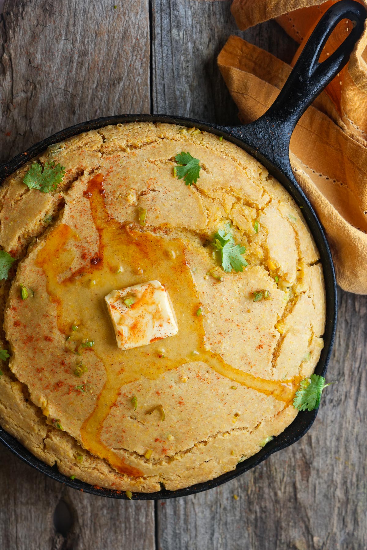 A cast iron skillet filled with vegan cornbread, garnished with a pat of butter and cilantro, placed on a rustic wooden surface next to a folded yellow cloth.