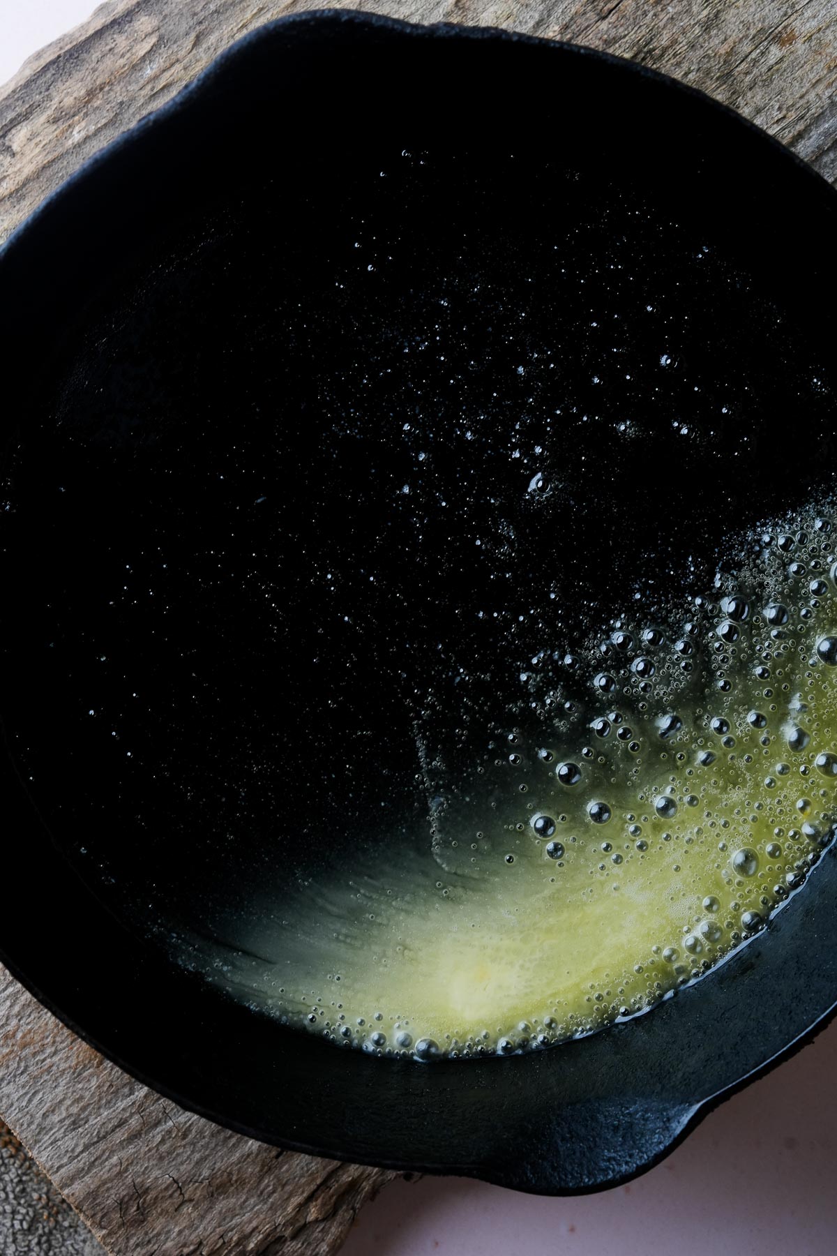 A cast iron skillet with melting vegan butter on a wooden surface, with bubbles forming along the edge of the melted area.