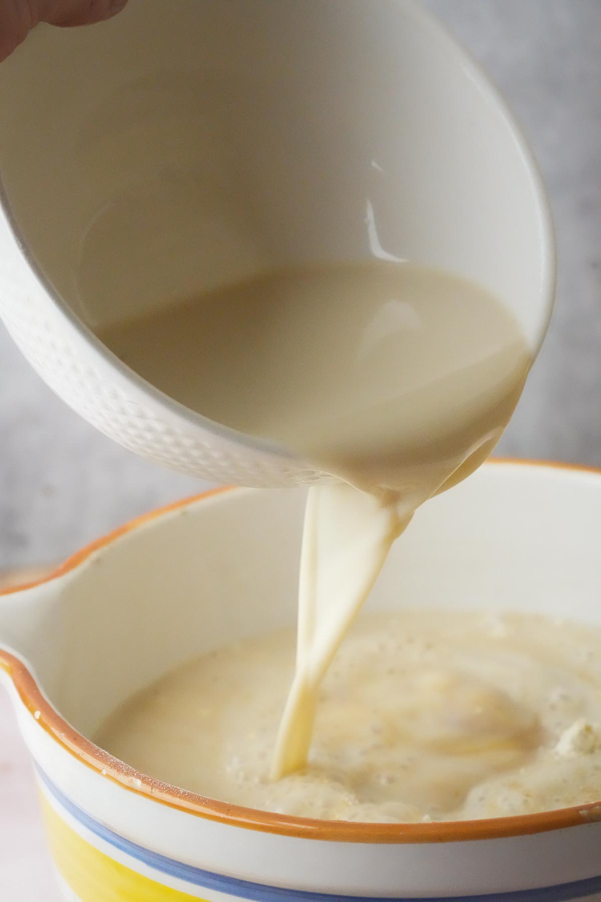 A white bowl with a yellow rim is pouring vegan milk into another bowl with a similar design.